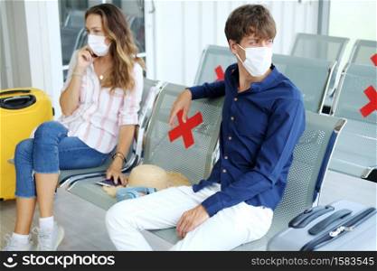 Serious Young Caucasian traveler couple love is wearing mask and sitting passenger waiting chairs for time of departure together because Plane delay in the airport. Honeymoon trip on vacation concept.