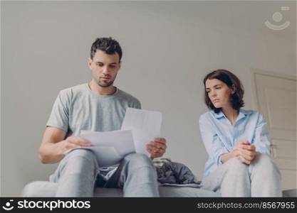 Serious young couple study documents together at bed, have serious looks, plan thier budget, pose against domestic interior, dressed in casual clothes. People, family and paperwork concept.