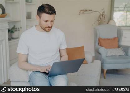 Serious young man using laptop computer for remote work from home, focused freelancer guy reading incoming emails or chatting online, researching or studying while sitting on couch in living room. Serious young man using laptop computer for remote work from home