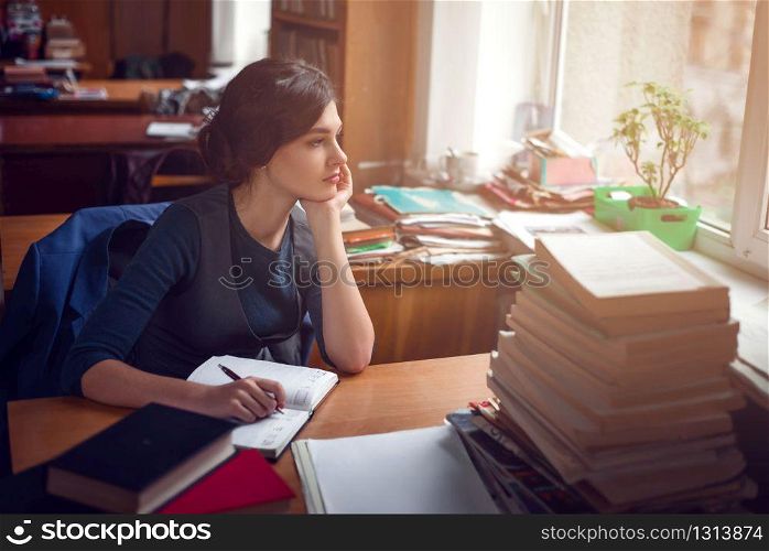 Serious young woman sitting at a table among books and thinking in library silence.