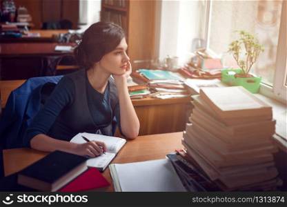 Serious young woman sitting at a table among books and thinking in library silence.