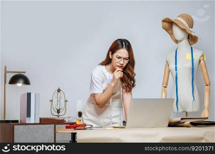 Seriously young Asian female fashion designer or costume use laptop computer while design a beautiful sombrero and taking on mannequin