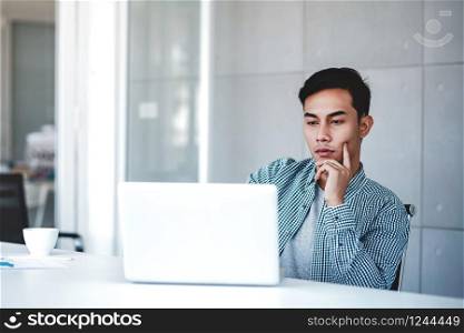 Seriously Young Businessman Working on Computer Laptop in Office. Hand on Shin, Sitting on Desk with Thoughtful Posture. Concentrated and Smart Men