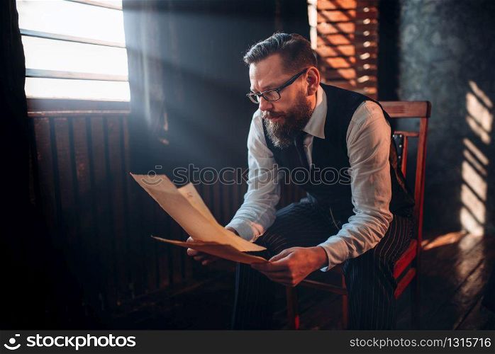 Seriuos bearded writer in glasses reads handwritten text against the window with sunlight. Retro typewriter, feather, crystal decanter, books and vintage lamp on the desk. Bearded writer in glasses reads handwritten text