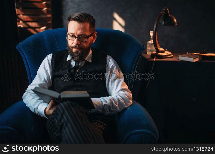 Seriuos bearded writer in glasses reads handwritten text against the window with sunlight. Retro typewriter, feather, crystal decanter, books and vintage lamp on the desk. Bearded writer in glasses reads handwritten text