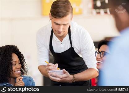 service and people concept - group of happy friends and waiter with notepad receiving order or restaurant. customers and waiter with notepad or restaurant