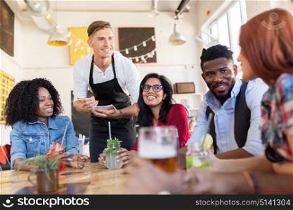 service and people concept - group of happy international friends with drinks and waiter with notepad receiving order at bar or restaurant. waiter and friends with menu and drinks at bar