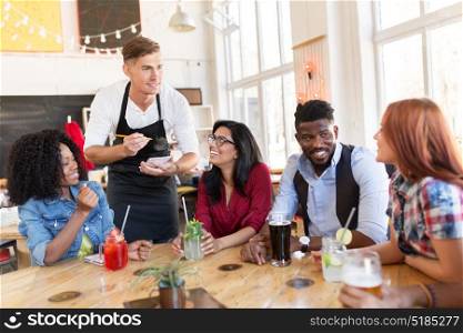 service and people concept - group of happy international friends with drinks and waiter with notepad receiving order at bar or restaurant. waiter and friends with menu and drinks at bar
