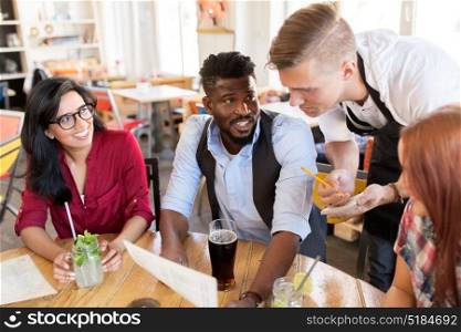service and people concept - group of happy international friends with menu and drinks and waiter with notepad receiving order at bar or restaurant. waiter and friends with menu and drinks at bar