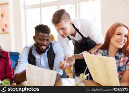 service and people concept - group of happy international friends with menu and drinks and waiter with notepad receiving order at bar or restaurant. waiter and friends with menu and drinks at bar