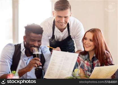 service and people concept - happy couple with menu and drinks and waiter with notepad receiving order at bar or restaurant. waiter and couple with menu and drinks at bar