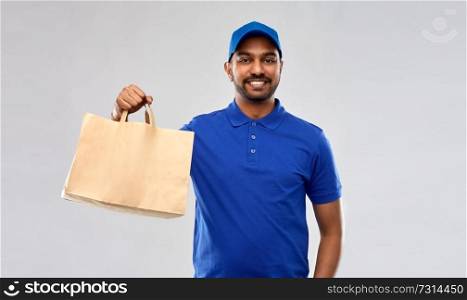 service and people concept - happy indian delivery man food in paper bag in blue uniform over grey background. happy indian delivery man with food in paper bag