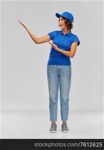 service and people concept - happy smiling delivery woman in blue uniform pointing to something imaginary over grey background. happy smiling delivery woman in blue uniform