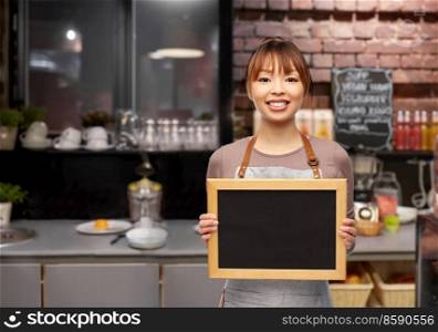 service and people concept - happy smiling female barista or waitress in apron with chalkboard over bar counter at restaurant background. happy female barista with chalkboard at restaurant