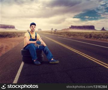 Service mechanic in uniform sitting on tire, desert road on background. Repairman, wheel mounting, tyre service