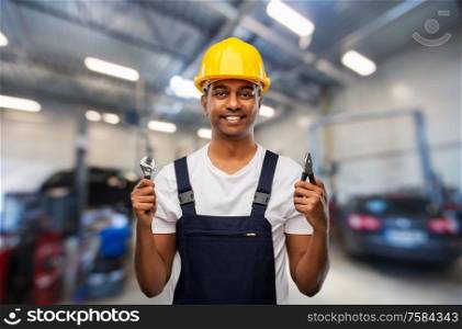 service, repair and profession concept - happy smiling indian mechanic in helmet with wrench and pliers over car shop background. indian mechanic with wrench and pliers at car shop