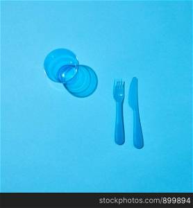 Set of disposable plastic utensil and empty glass with shadows on a pastel blue background, copy space. Top view. Plastic glass, fork and knife with shadows on a blue table.