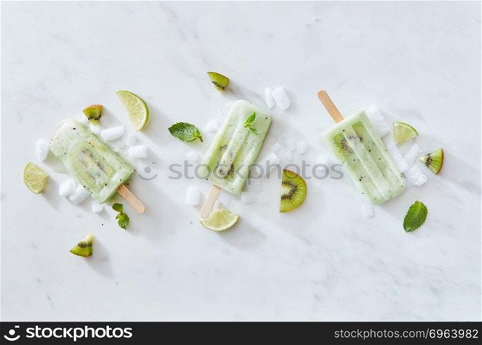 Set of ice cream on a stick with kiwi, mint and lime on a gray marble background with pieces of ice and fruit. Space for text. Flat lay. Sticks of ice cream with kiwi and lime on a gray marble background with pieces of ice and mint leaves. Copy space. Flat lay