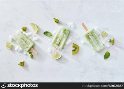 Set of ice cream on a stick with kiwi, mint and lime on a gray marble background with pieces of ice and fruit. Space for text. Flat lay. Sticks of ice cream with kiwi and lime on a gray marble background with pieces of ice and mint leaves. Copy space. Flat lay