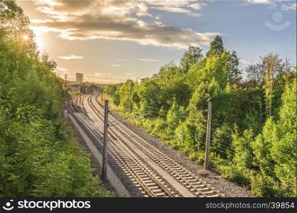 Set of railways going towards a train station, surrounded by green trees and under the warm light of a sun setting down.