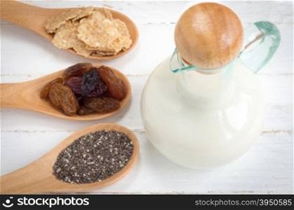 Set of raisins, whole wheat grain flakes and chia seeds in wooden spoon and milk on white wood background&#xA;