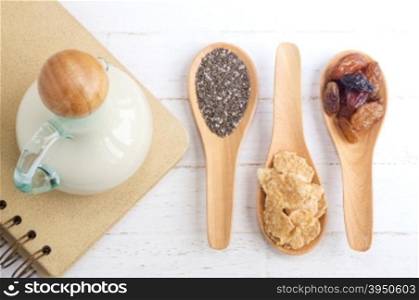 Set of raisins, whole wheat grain flakes and chia seeds in wooden spoon and milk on white wood background&#xA;