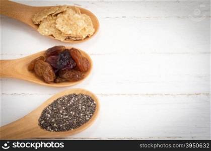 Set of raisins, whole wheat grain flakes and chia seeds in wooden spoon on white wood background.