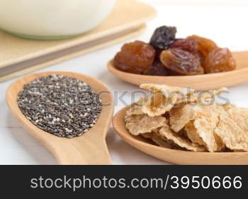 Set of raisins, whole wheat grain flakes and chia seeds in wooden spoon and milk on white wood background with selective focus point&#xA;