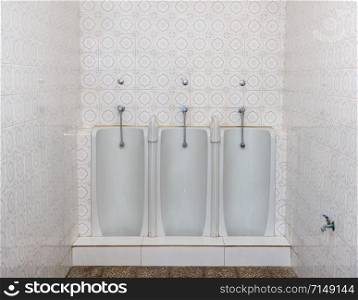 Set of three side by side full length mens urinals in porcelain against a tiled wall in small restroom. Three full length urinals in a row on a tiled bathroom or restroom wall