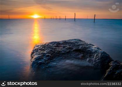 Setting sun above Dutch IJsselmeer with boulders in foreground, Lake IJssel, Flevoland, Netherlands. Setting sun al lake with fishing net poles and rocky coast