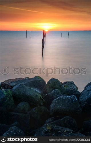Setting sun above Dutch IJsselmeer with boulders in foreground, Lake IJssel, Flevoland, Netherlands. Sundown with fish trap at the coast