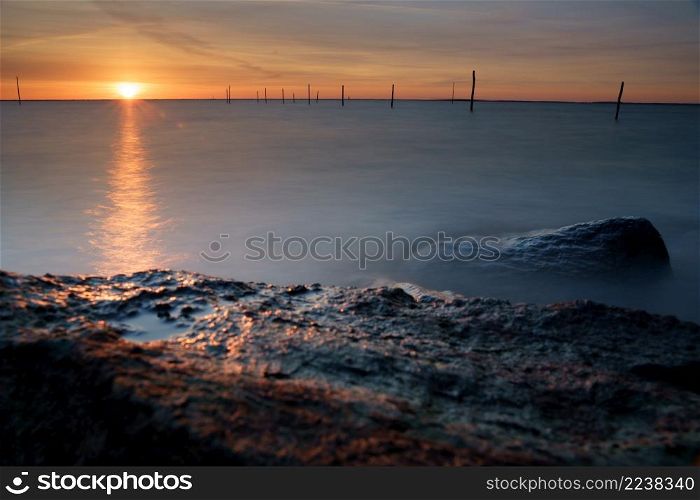 Setting sun above Dutch IJsselmeer with boulders in foreground, Lake IJssel, Flevoland, Netherlands. Setting sun al lake with fishing net poles and rocky coast