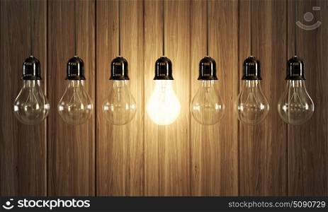 Seven light bulbs with glowing one on wooden background.
