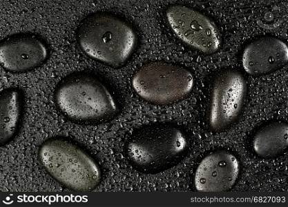 Several black basalt massage stones, covered with water drops, distributed on a black background; top view, flat lay