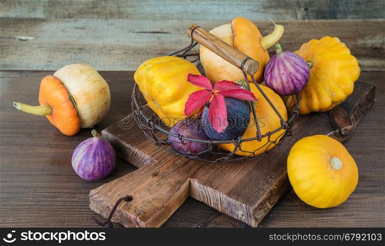 Several orange decorative pumpkins and patissons and blue figs are in a mesh metal basket on a dark wooden background