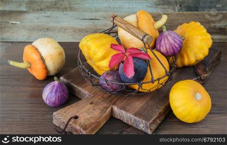 Several orange decorative pumpkins and patissons and blue figs are in a mesh metal basket on a dark wooden background