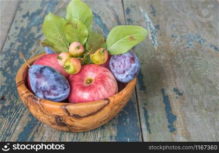 Several red apples with green leaves, blue plums, pear and rowan lie in a wooden bowl on the old wooden table, with copy-space