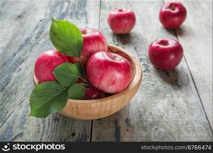 Several red apples with green leaves lie in a wooden bowl on the old wooden table