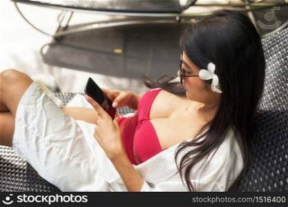 sexy Asian beautiful woman in red swimsuit and sunglasses play social media, chat, shop online by smartphone on bed chair of swiming pool. Tan girl summer vacation at luxury hotel resort.