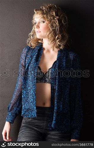 sexy blond curly haired woman in blue shirt and bra against a black wall