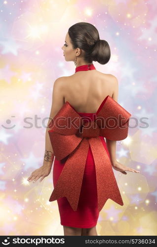 sexy brunette girl with elegant christmas style posing turned on her back with elegant red dress with big glitter bow, naked shoulders and golden jewellery