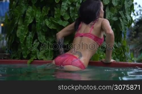 Sexy brunette woman wearing pink bikini relaxing at the edge of swimming pool. Young female is watching beautiful view and enjoying her vacation at tropical resort - slow motion video