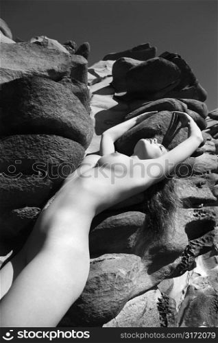 Sexy Caucasian young adult woman with eyes closed standing nude against rocks at Maui coast with arms stretched over head.