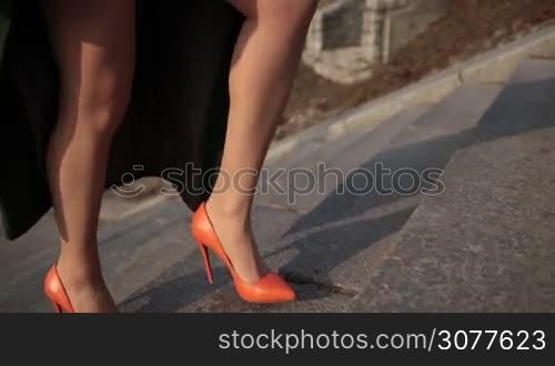 Sexy female legs in orange high heels shoes going up stone stairs in city. Elegant woman wearing mini skirt and emerald green coat walking up the stairs outdoors. Closeup beautiful female legs in high heels stepping up. Angle view. Slow motion.