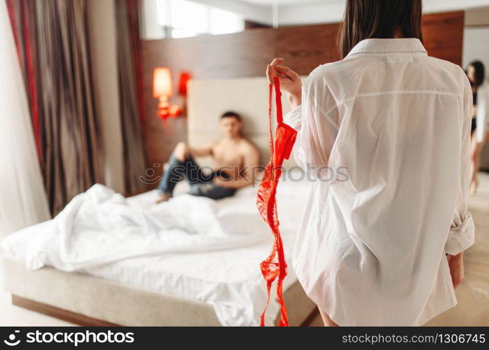 Sexy female person with red erotic bra in hand, man lies on big white bed. Intimate couple in bedroom, sex lovers. Sexy female with red bra in hand, itimate couple