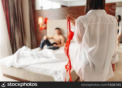 Sexy female person with red erotic bra in hand, man lies on big white bed. Intimate couple in bedroom, sex lovers. Sexy female with red bra in hand, itimate couple