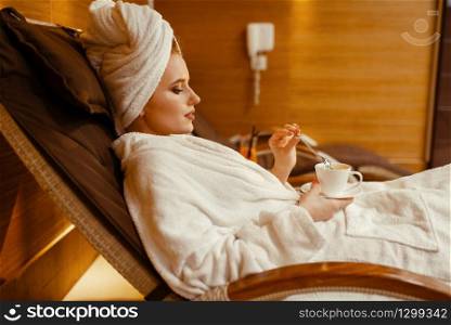Sexy girl in bathrobe and towel on the head relaxing with cup of coffee in spa chair. Relaxation leisure, healthy lifestyle, attractive woman resting in armchair, beauty salon. Sexy girl relaxing with cup of coffee in spa chair