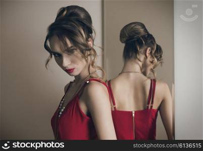 sexy girl with angry expression posing in changing room near mirror with red dress, elegant hair-style and make-up.
