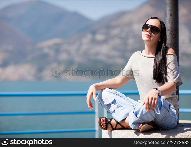 sexy girl with sunglasses is sitting and posing in front of a water background