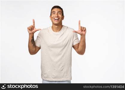 Sexy hispanic muscular male in casual t-shirt, looking up joyful, pointing top advertisement, laughing and smiling from delight and joy, standing white background amused, see something beautiful.. Sexy hispanic muscular male in casual t-shirt, looking up joyful, pointing top advertisement, laughing and smiling from delight and joy, standing white background amused, see something beautiful